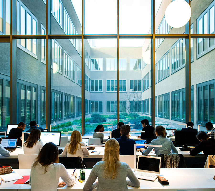 Viele Studierende arbeiten an Computern in einem modernen Lernraum mit Blick durch eine große Glasfront auf einen Innenhof.