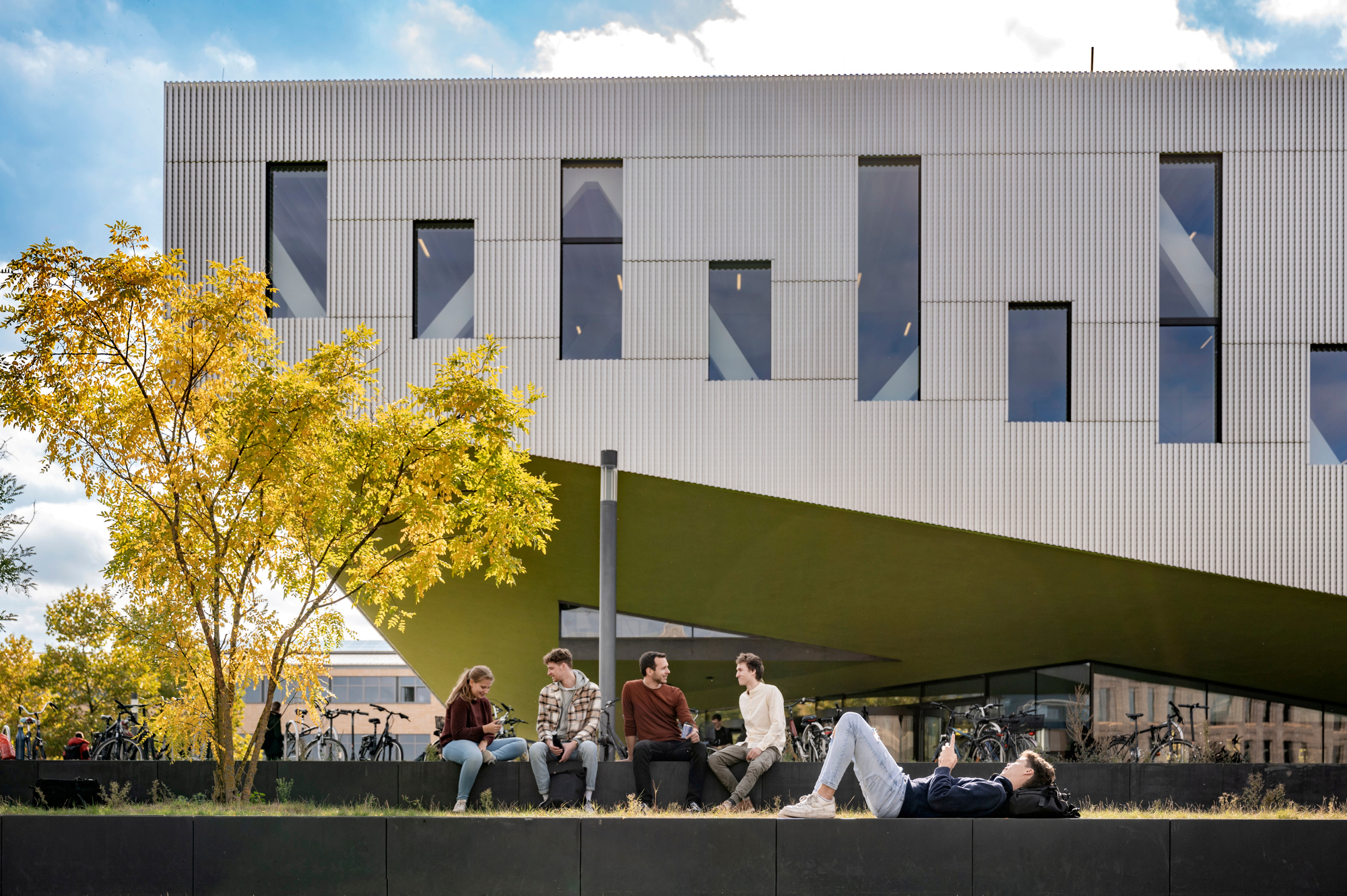 Fünf Studierende entspannen vor einem modernen Gebäude der Hochschule Osnabrück. Ein Student liegt auf einer Mauer, die anderen sitzen daneben.