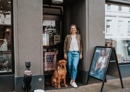 Lächelnde Frau steht mit einem braunen Hund im Eingang des Geschäfts Hundeklunker. Daneben steht eine schwarze Hundestatue.