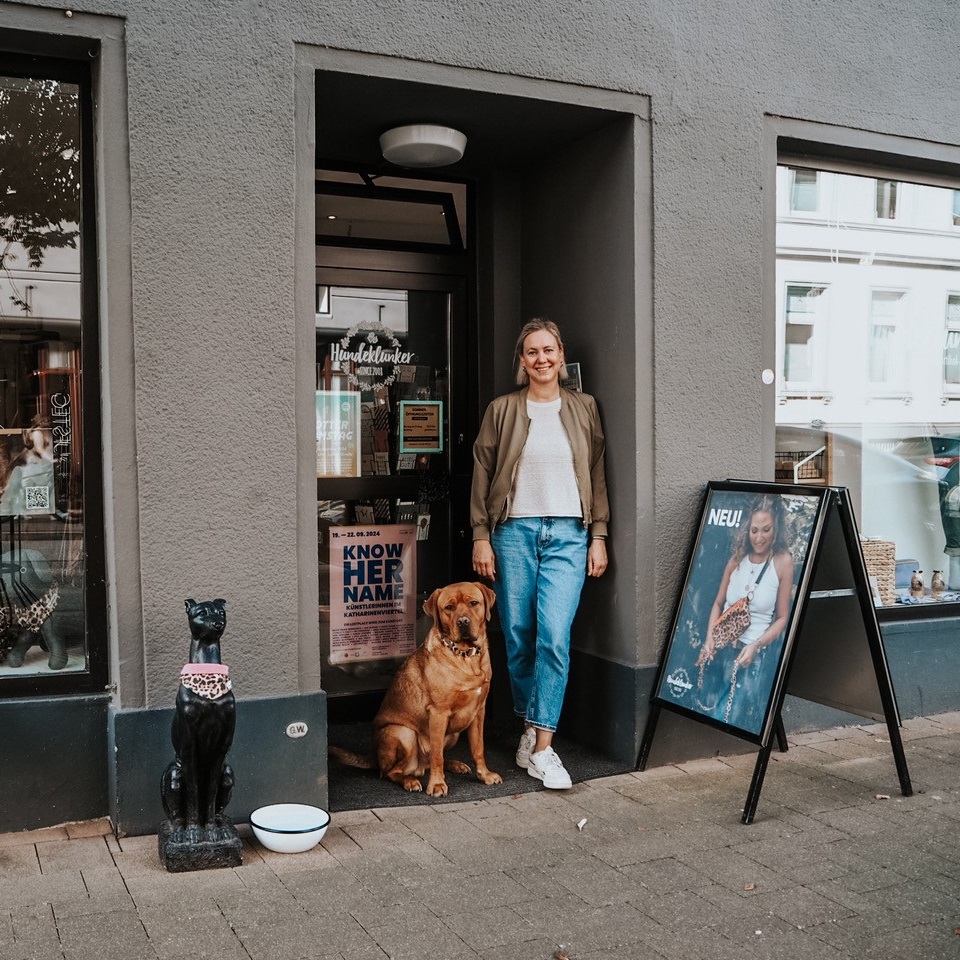 Lächelnde Frau steht mit einem braunen Hund im Eingang des Geschäfts Hundeklunker. Daneben steht eine schwarze Hundestatue.