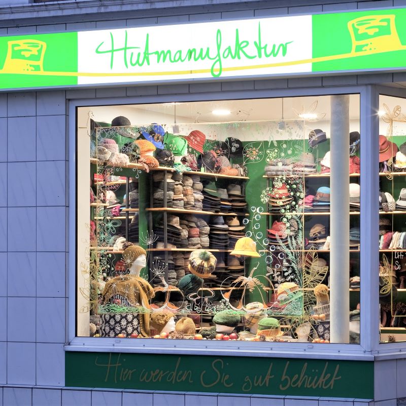 Schaufenster eines Hutgeschäfts mit dem Schriftzug Hutmanufaktur. Im Regal sind zahlreiche Hüte aufgereiht, unten steht: Hier werden Sie gut behütet.