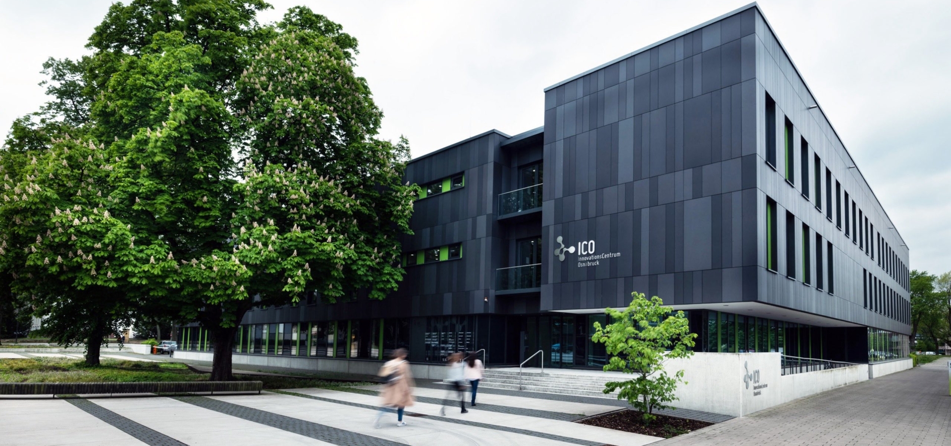 Moderne dunkle Gebäudefassade des InnovationCentrums Osnabrück mit großen Fensterfronten und gepflastertem Vorplatz.