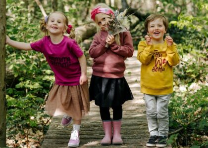 Drei fröhliche Kinder stehen auf einem Holzsteg im Wald. Das linke Mädchen trägt ein pinkes Shirt mit dem Schriftzug „REMMIDEMMI“