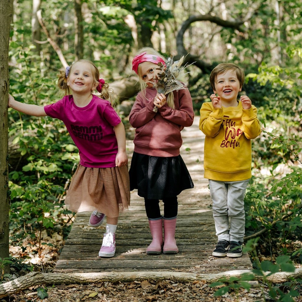 Drei fröhliche Kinder stehen auf einem Holzsteg im Wald. Das linke Mädchen trägt ein pinkes Shirt mit dem Schriftzug „REMMIDEMMI“