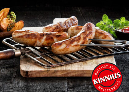 Gegrillte Bratwürste auf einem Grillrost über Holzbrett, mit Logo KINNIUS – Qualität aus Meisterhand und Schriftzug: Die Meister-Wurst.