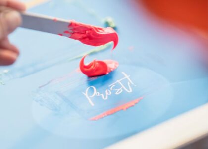 Pinkfarbene Farbe wird mit einem Spachtel auf ein Siebdrucksieb aufgetragen. Darunter befindet sich der Schriftzug: Prost!