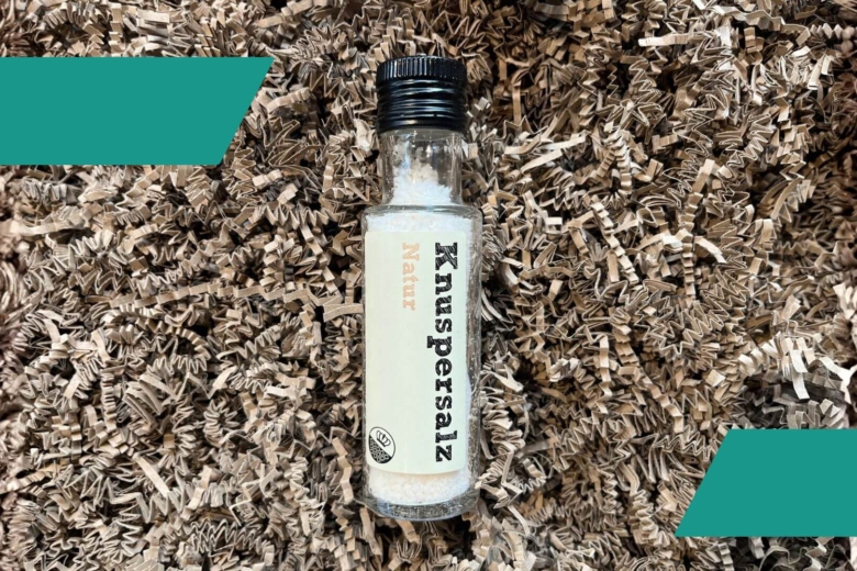 Schlanke Glasflasche mit der Aufschrift Knuspersalz Natur auf braunem Verpackungsmaterial.