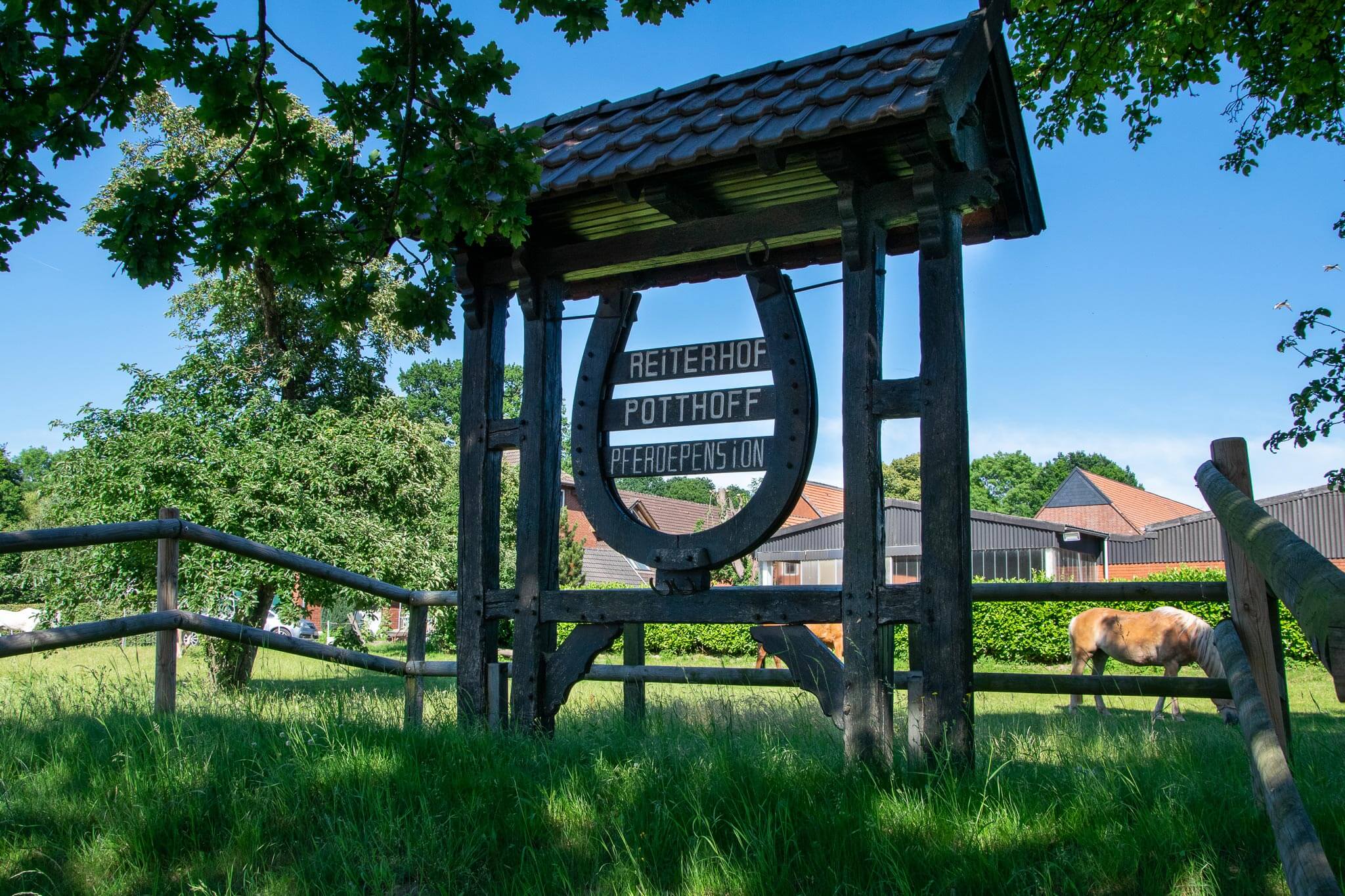 Rustikaler Holzbogen mit der Aufschrift „Reiterhof Potthoff“ vor einer Koppel mit grasendem Pferd.