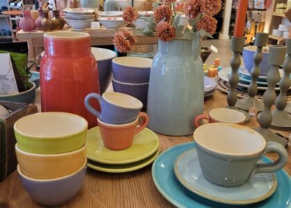 Buntes Keramikgeschirr, Schalen, Tassen und eine hellblaue Vase mit Trockenblumen stehen auf einem Holztisch in einem Dekorationsgeschäft.