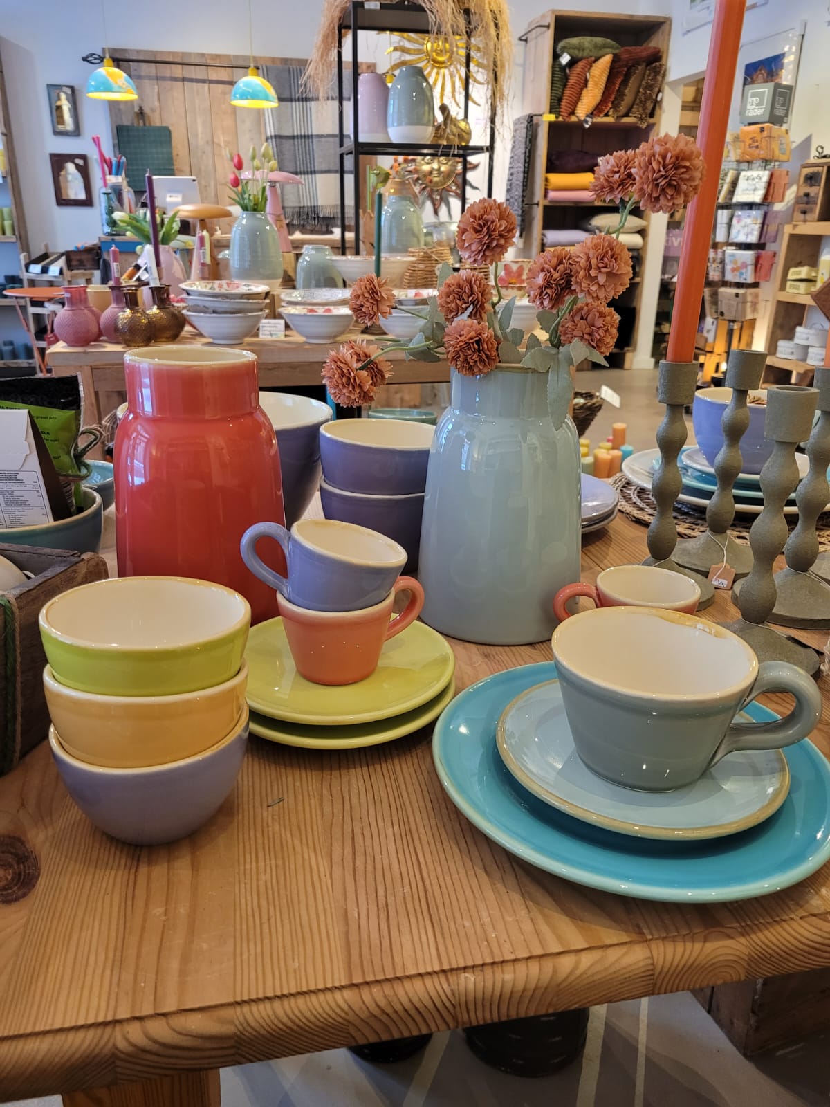 Buntes Keramikgeschirr, Schalen, Tassen und eine hellblaue Vase mit Trockenblumen stehen auf einem Holztisch in einem Dekorationsgeschäft.