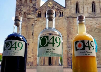 Drei O49-Spirituosenflaschen (Organic Amaro, Gin, Advocaat) stehen nebeneinander auf einer Mauer vor der Kulisse einer historischen Stein-Kirche.