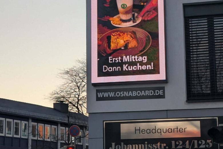 Eine vertikale digitale Werbefläche an einer Hauswand zeigt Gastronomiewerbung für die Portobar mit Kaffee und Kuchen.