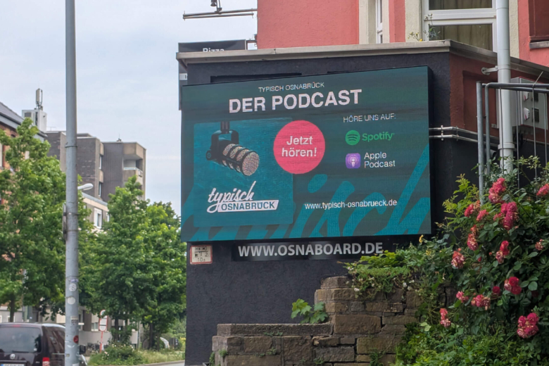 Ein digitales Werbeschild an einem Gebäude wirbt für den Podcast Typisch Osnabrück mit Verweisen zu Streaming-Diensten.