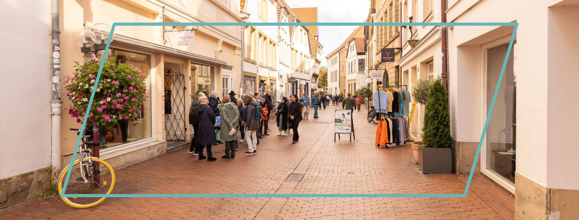 Fußgänger schlendern durch eine malerische Altstadtgasse mit historischen Fachwerkhäusern, Kopfsteinpflaster und blühenden Blumenampeln.
