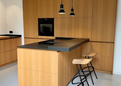 Moderne Küche aus hellem Holz mit Kochinsel, schwarzer Arbeitsplatte, zwei Barhockern und zwei hängenden Lampen vor Einbauschränken mit Backofen.