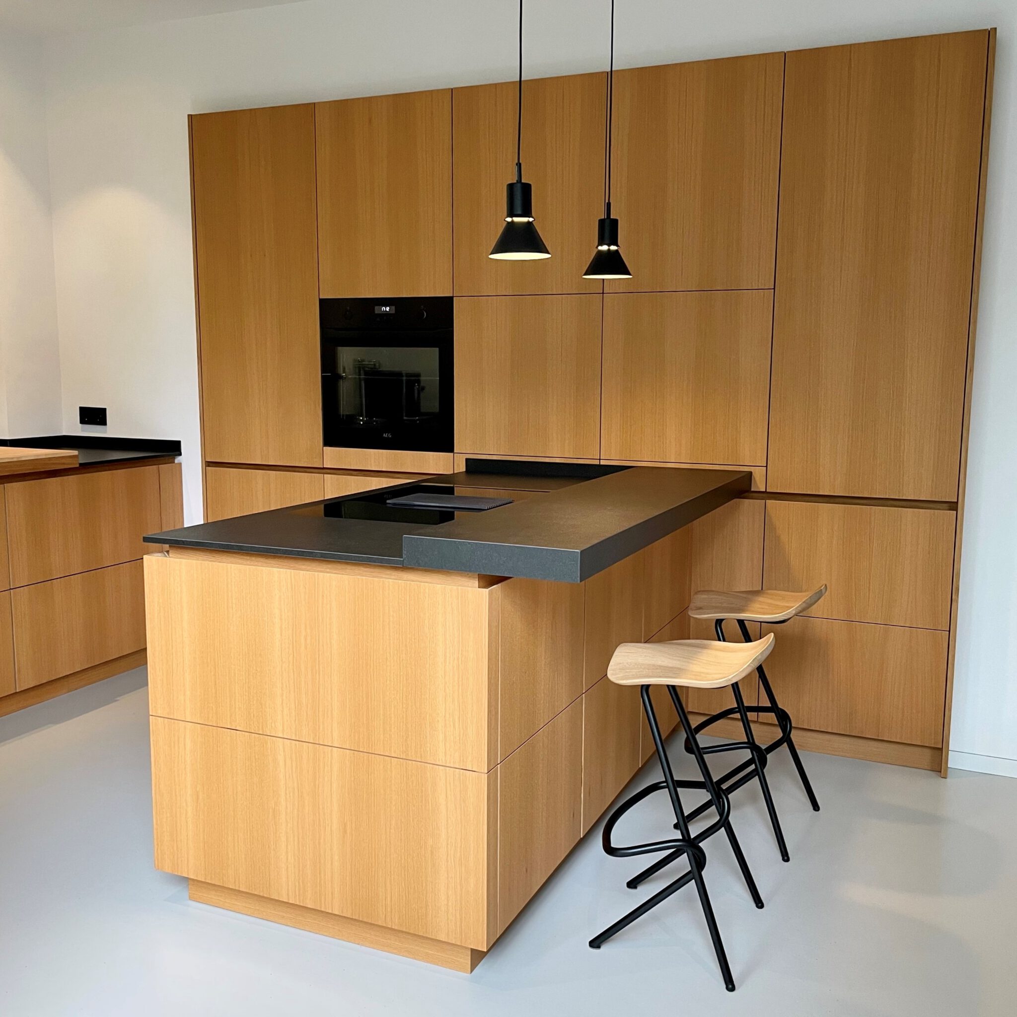 Moderne Küche aus hellem Holz mit Kochinsel, schwarzer Arbeitsplatte, zwei Barhockern und zwei hängenden Lampen vor Einbauschränken mit Backofen.
