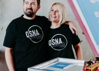 Ein lächelndes Paar steht an einem Siebdruckrahmen und trägt schwarze T-Shirts mit dem kreisförmigen Aufdruck: OSNA merch.com.