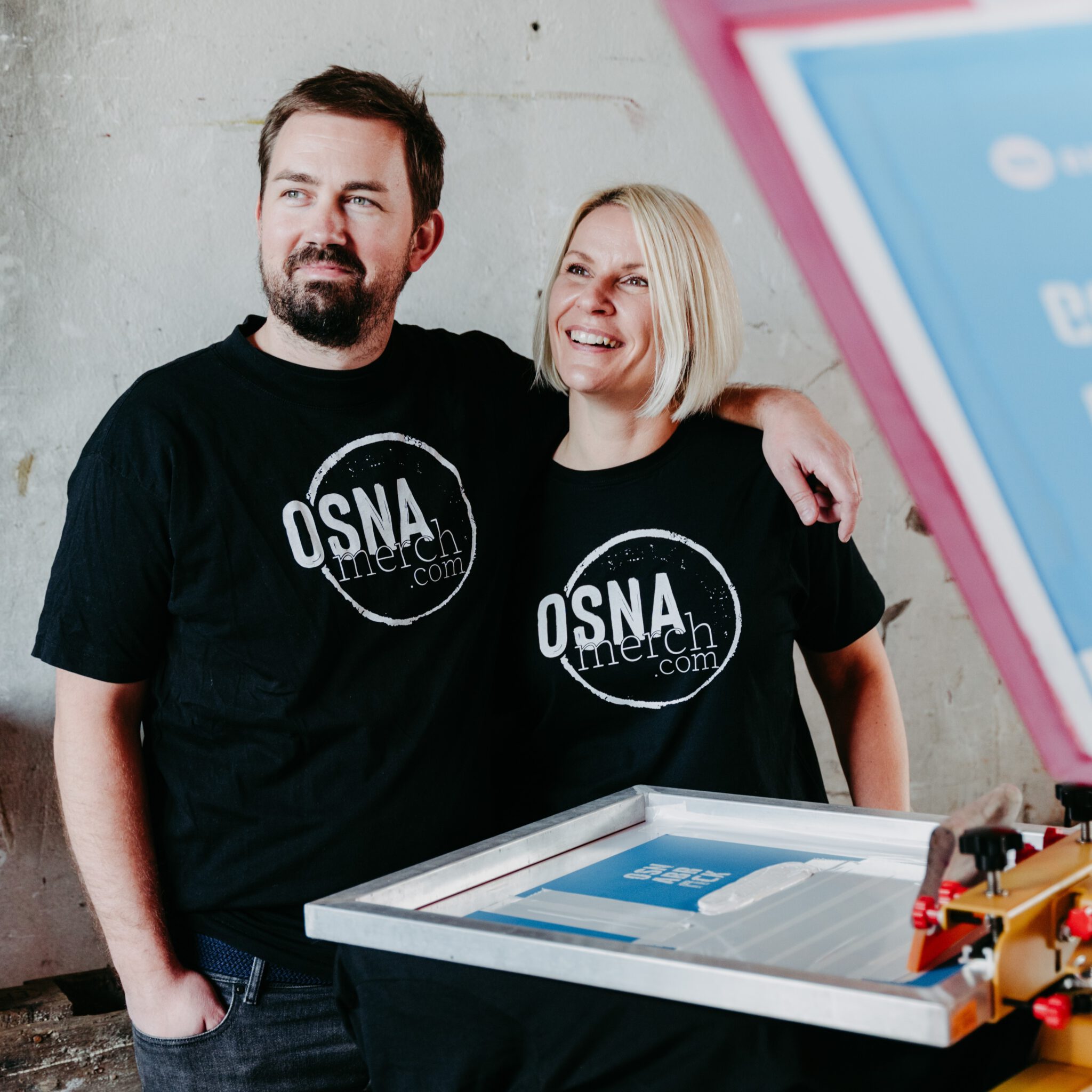 Ein lächelndes Paar steht an einem Siebdruckrahmen und trägt schwarze T-Shirts mit dem kreisförmigen Aufdruck: OSNA merch.com.