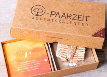 Geöffneter „Paarzeit Adventskalender“ aus Braunpappe mit Kärtchen und Holzwäscheklammern. Auf Karte 1 steht: „Verliebtsein ist, wenn…“
