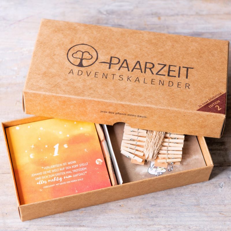 Geöffneter „Paarzeit Adventskalender“ aus Braunpappe mit Kärtchen und Holzwäscheklammern. Auf Karte 1 steht: „Verliebtsein ist, wenn…“