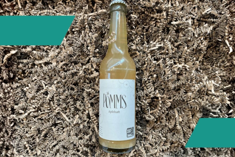 Glasflasche mit naturtrübem Apfelschaumwein und weißem Pömms-Etikett auf braunem Füllmaterial.