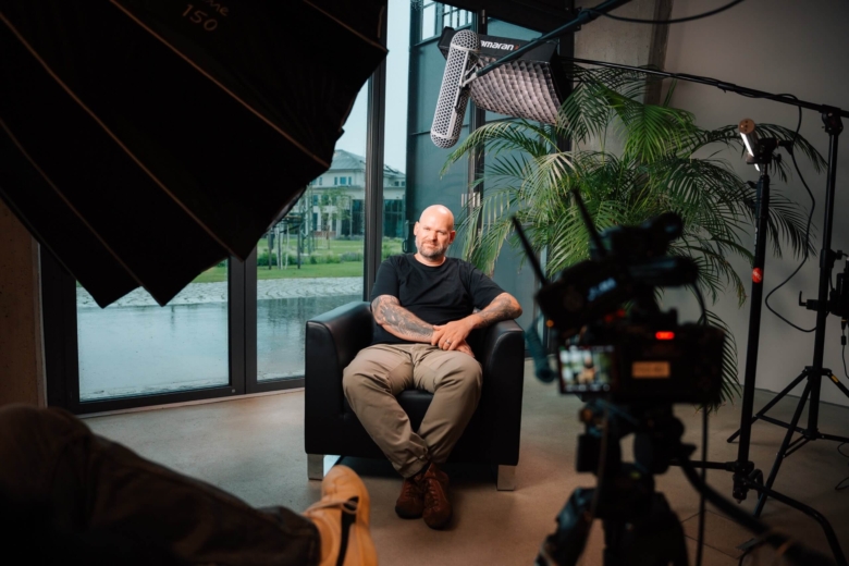 Tätowierter Mann sitzt entspannt in einem Sessel vor Kamera und Film-Equipment im Studio.