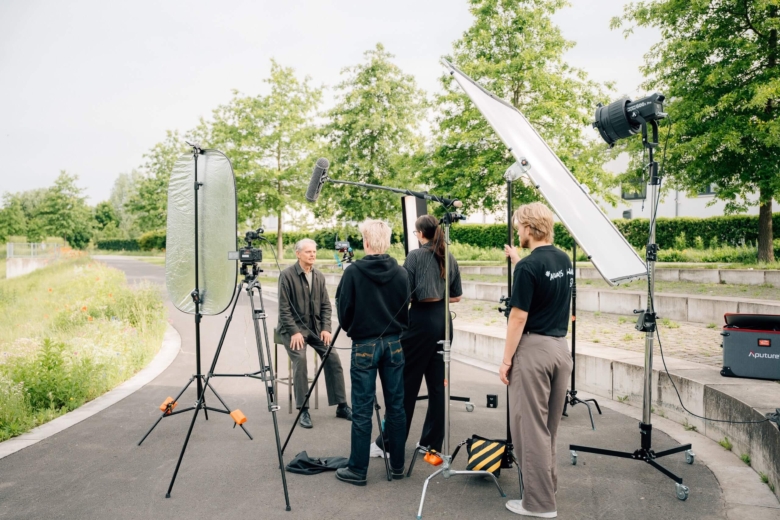 Aufnahmeteam filmt einen sitzenden Mann im Freien mit Kamera, Mikrofon und Licht-Reflektoren.