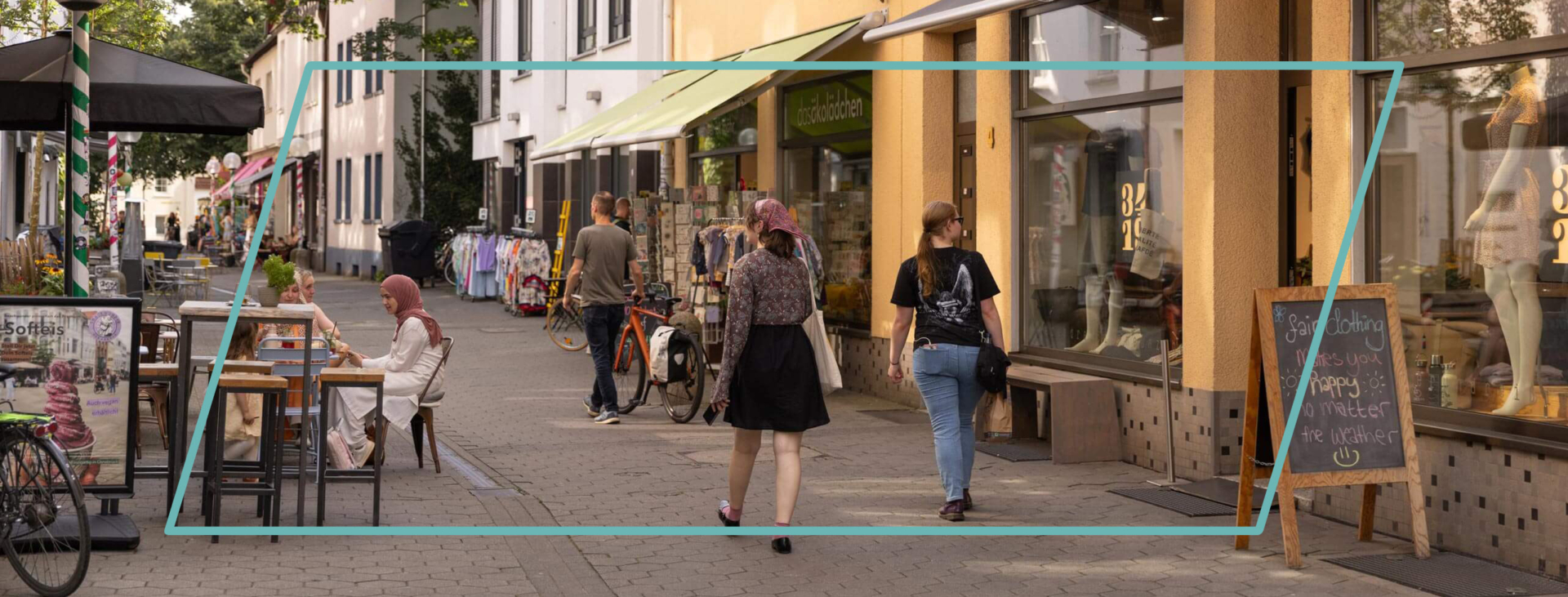 Menschen flanieren durch eine sonnige Einkaufsstraße mit gelben historischen Gebäuden, Straßencafés und gemütlichen Sitzgelegenheiten.