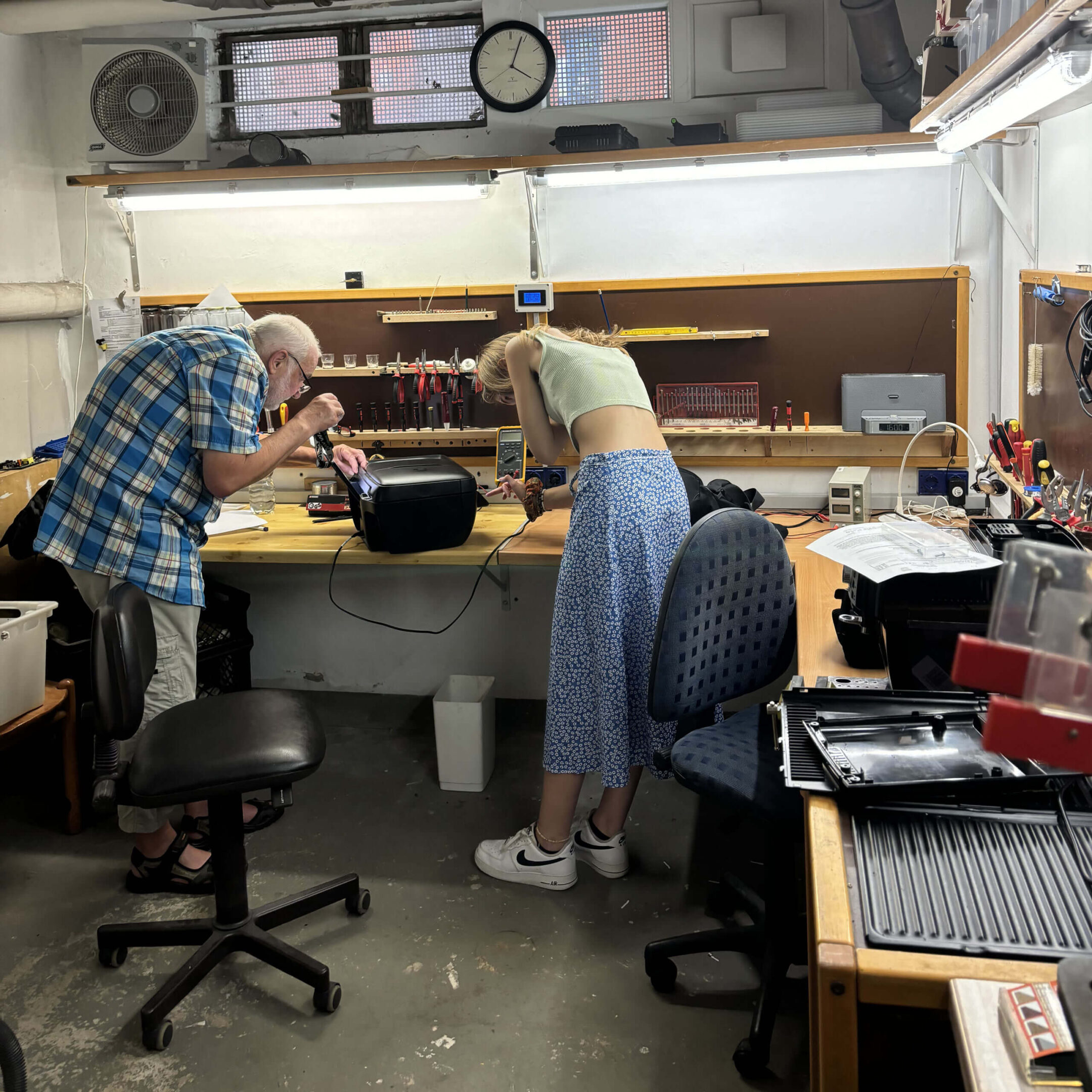 Eine Frau und ein älterer Mann untersuchen ein geöffnetes Elektronikgerät in einer Werkstatt.