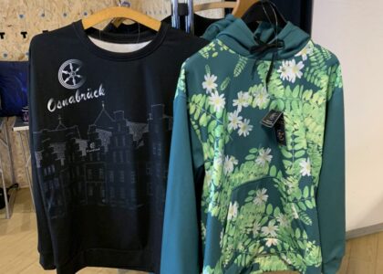Zwei bedruckte Pullover hängen an einem Ständer: links ein schwarzer mit Osnabrück-Motiv, rechts ein grüner Hoodie mit Blumenmuster.