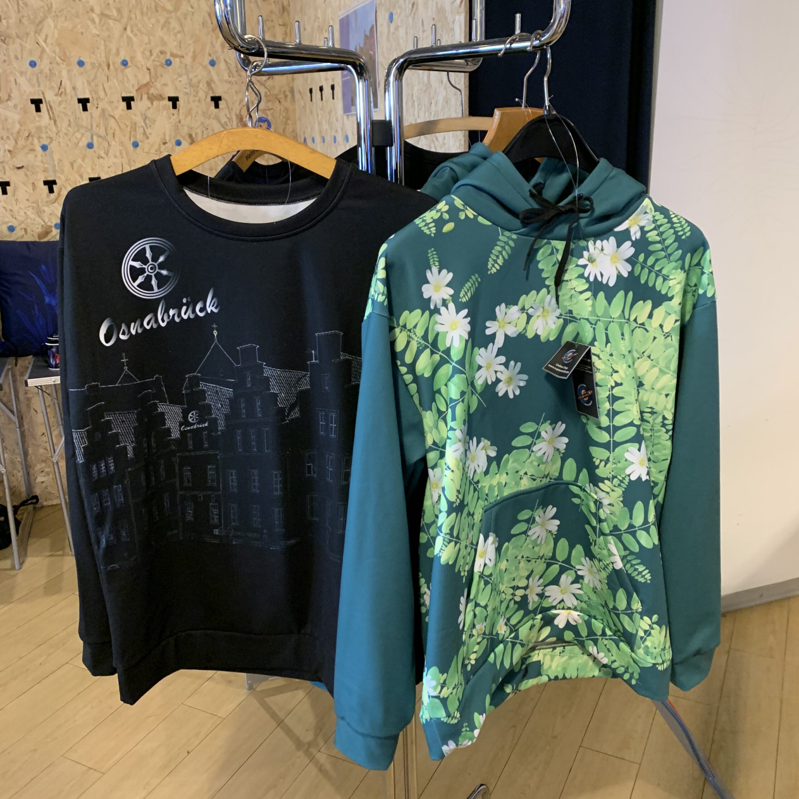 Zwei bedruckte Pullover hängen an einem Ständer: links ein schwarzer mit Osnabrück-Motiv, rechts ein grüner Hoodie mit Blumenmuster.