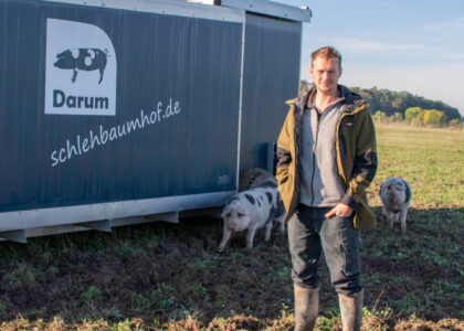 Landwirt auf einem Feld neben zwei Schweinen und einem Anhänger mit dem Schriftzug: Darum schlehbaumhof.de.