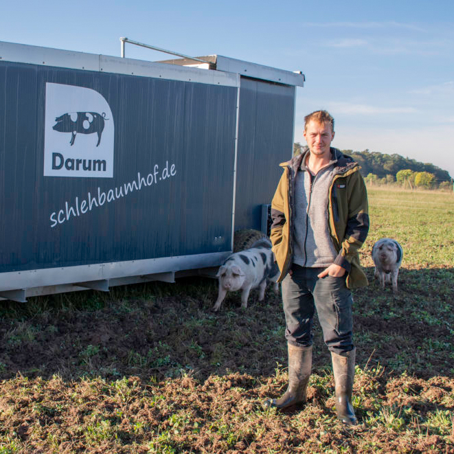 Landwirt auf einem Feld neben zwei Schweinen und einem Anhänger mit dem Schriftzug: Darum schlehbaumhof.de.