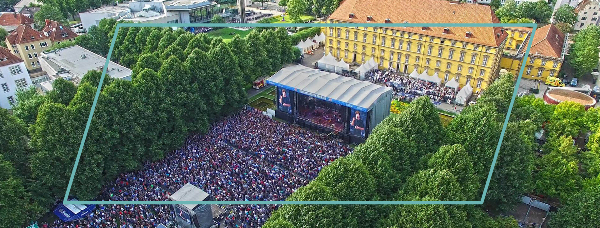Große Menschenmenge bei einem Open-Air-Konzert vor einer Hauptbühne im Schlossgarten Osnabrück, umgeben von Bäumen.