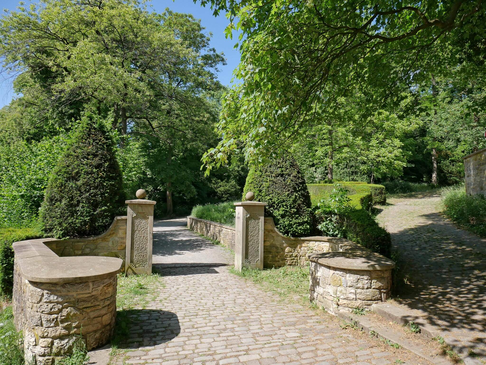 Gepflasterter Parkeingang mit Steinmauern und Posten führt in einen schattigen, grünen Park.