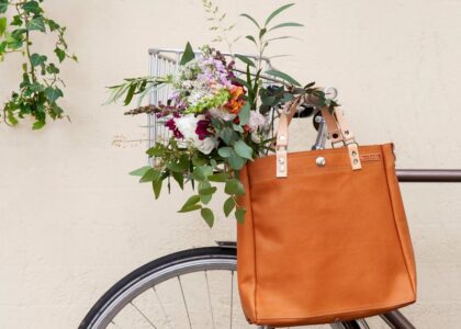 Braunfarbene Tasche und ein bunter Blumenstrauß in einem Drahtkorb an einem Fahrradlenker vor einer hellen Wand.