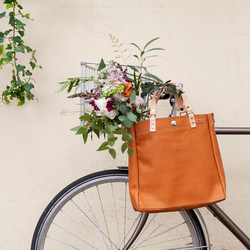 Braunfarbene Tasche und ein bunter Blumenstrauß in einem Drahtkorb an einem Fahrradlenker vor einer hellen Wand.