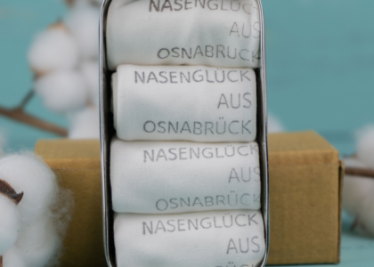 Vier gefaltete weiße Stofftaschentücher in Metalldose mit dem Aufdruck: NASENGLÜC"Nasenglück aus Osnabrück", vor Baumwollzweig.