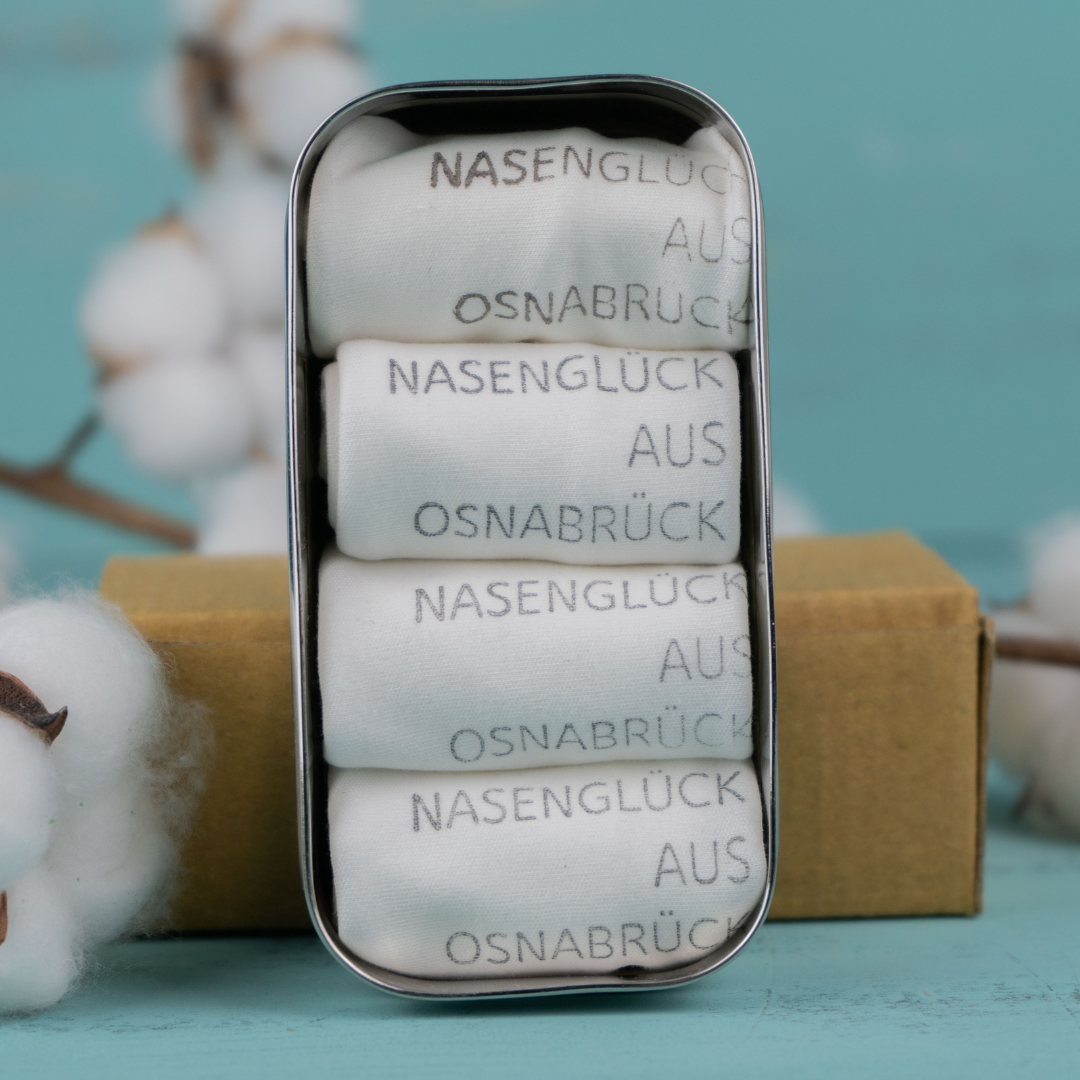 Vier gefaltete weiße Stofftaschentücher in Metalldose mit dem Aufdruck: NASENGLÜC"Nasenglück aus Osnabrück", vor Baumwollzweig.
