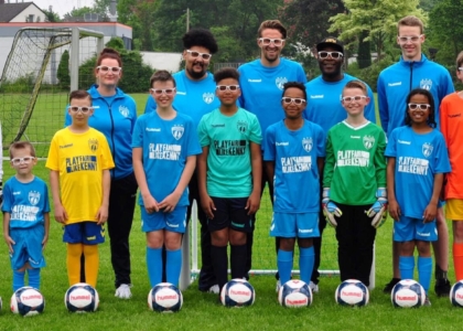 Jugend-Fußballmannschaft und Trainer in bunten Trikots und Brillen stehen nebeneinander auf dem Platz.