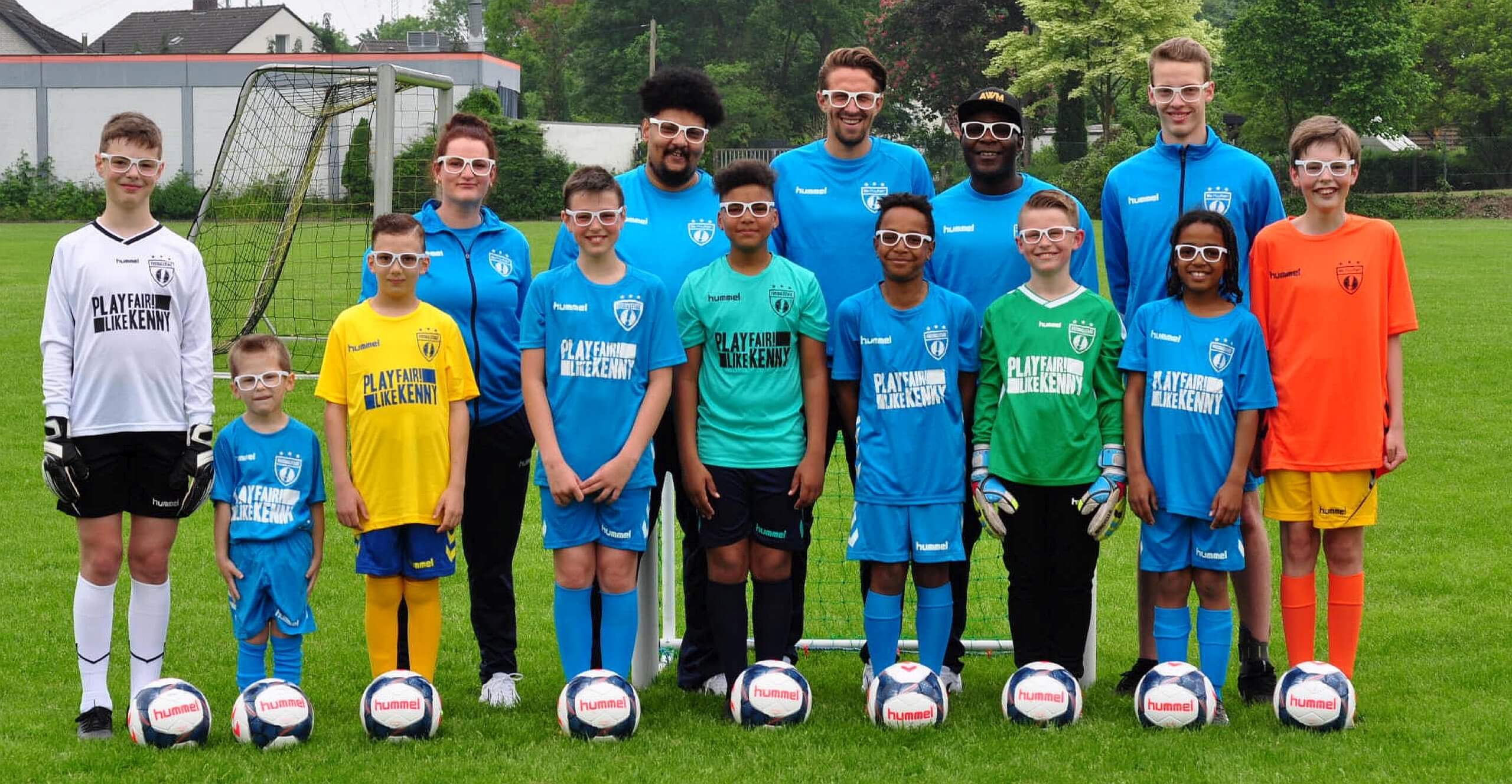 Jugend-Fußballmannschaft und Trainer in bunten Trikots und Brillen stehen nebeneinander auf dem Platz.
