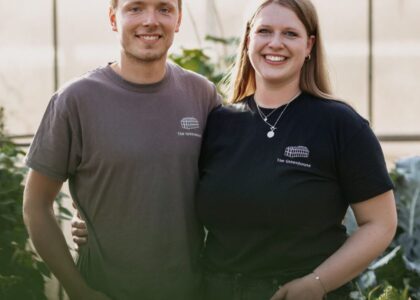 Lächelndes Paar im Gewächshaus in T-Shirts mit folgender Aufschrift: The Greenhouse. Sie halten einander umschlungen und blicken in die Kamera.