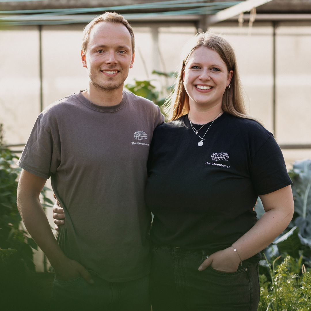 Lächelns Paar im Gewächshaus in T-Shirts mit folgender Aufschrift: The Greenhouse. Sie halten einander umschlungen und blicken in die Kamera.