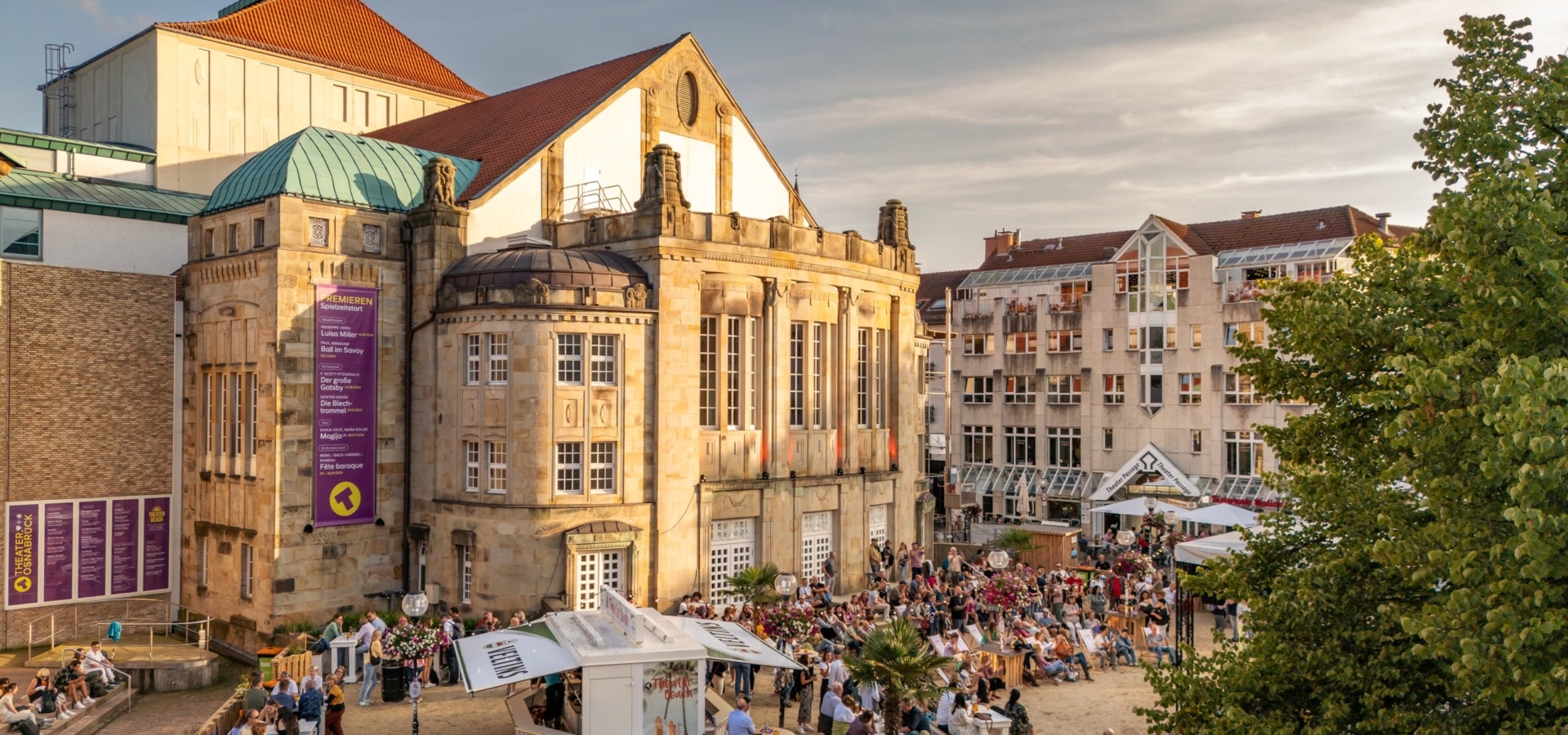 Menschenmenge genießt ein sommerliches Outdoor-Event mit Verkaufsständen vor dem historischen Theatergebäude.