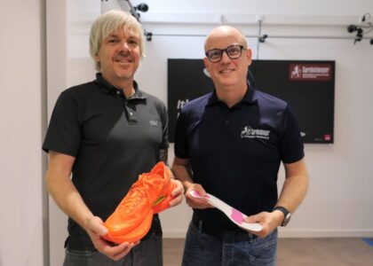 Zwei Männer präsentieren lächelnd einen orangefarbenen Laufschuh und eine Schuheinlage.
