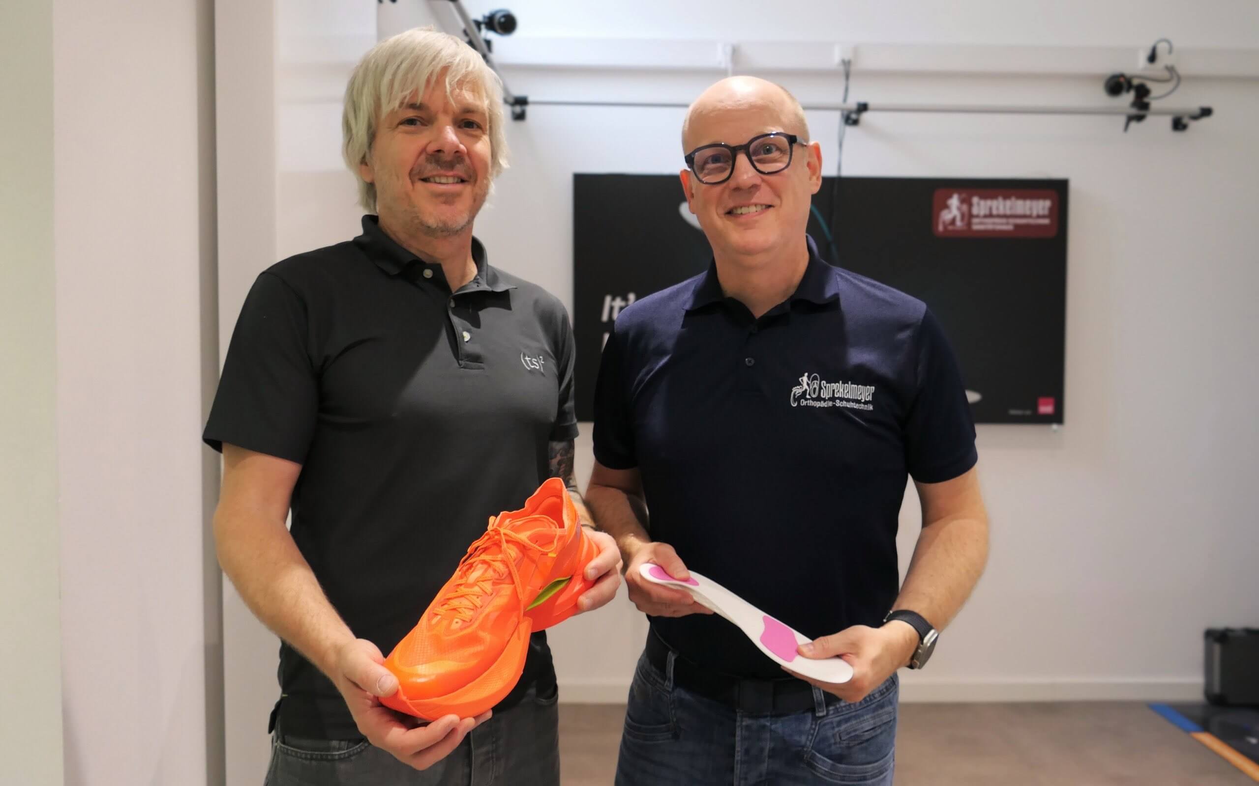 Zwei Männer präsentieren lächelnd einen orangefarbenen Laufschuh und eine Schuheinlage.