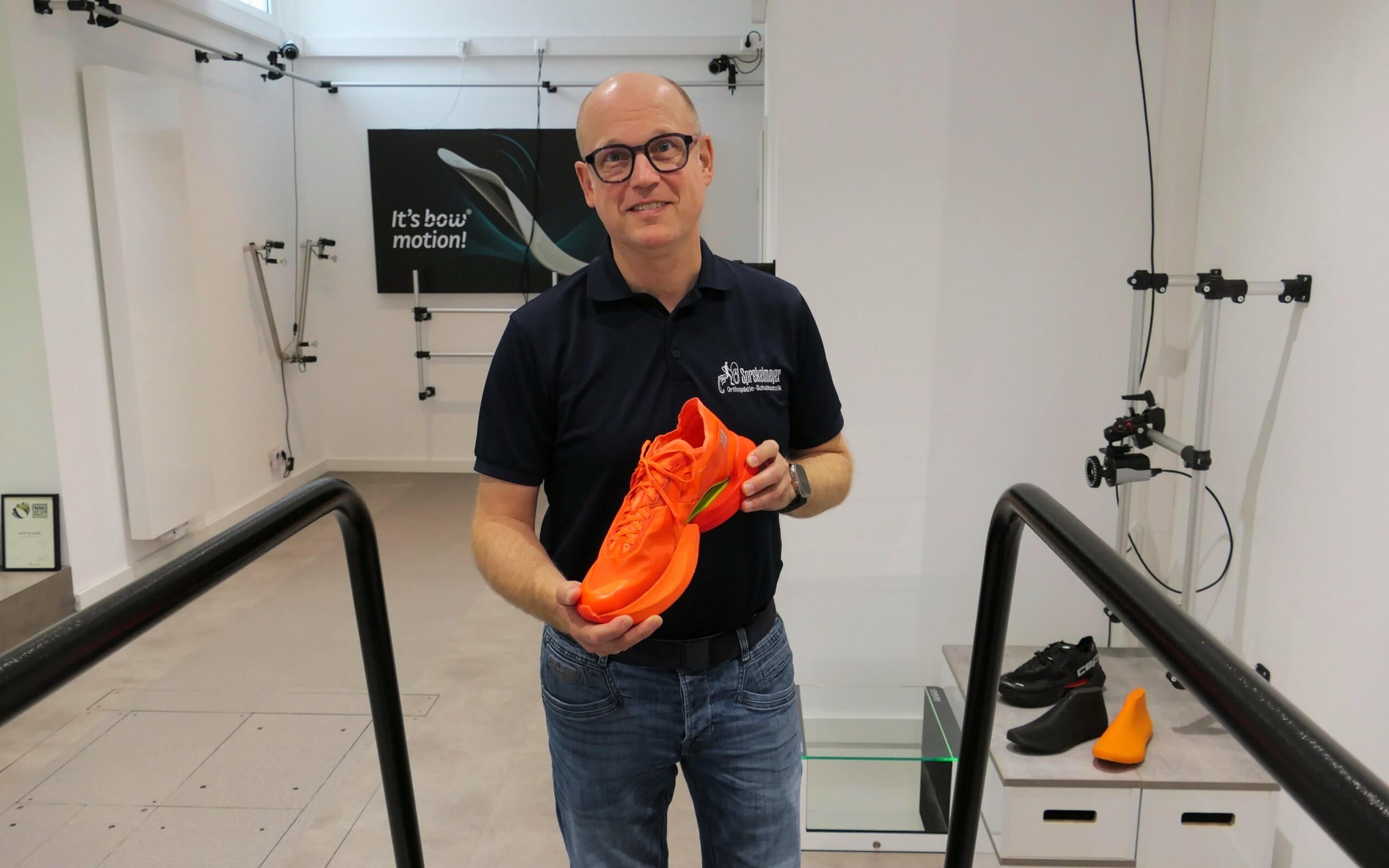 Mann steht mit Brille im Bewegungslabor und hält einen orangefarbenen Laufschuh.