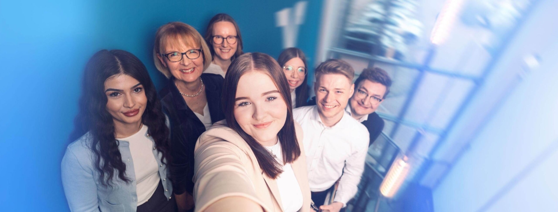 Eine Gruppe von sieben lächelnden Kolleginnen und Kollegen macht ein gemeinsames Selfie im Treppenhaus.