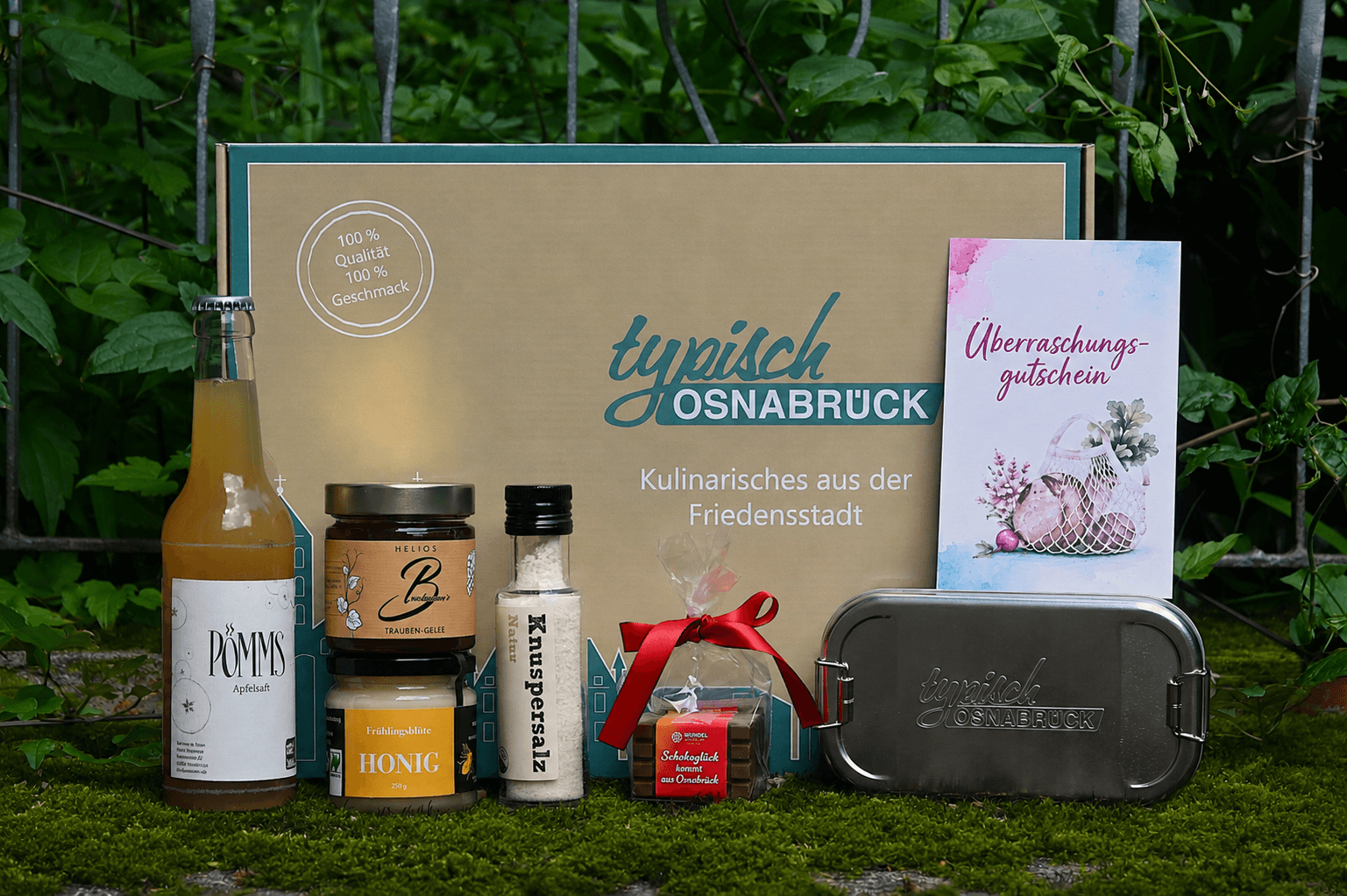 Geschenkbox „Typisch Osnabrück“ präsentiert mit Apfelsaft, Honig, Gelee, Salz, Schokolade und Edelstahl-Dose im Grünen.