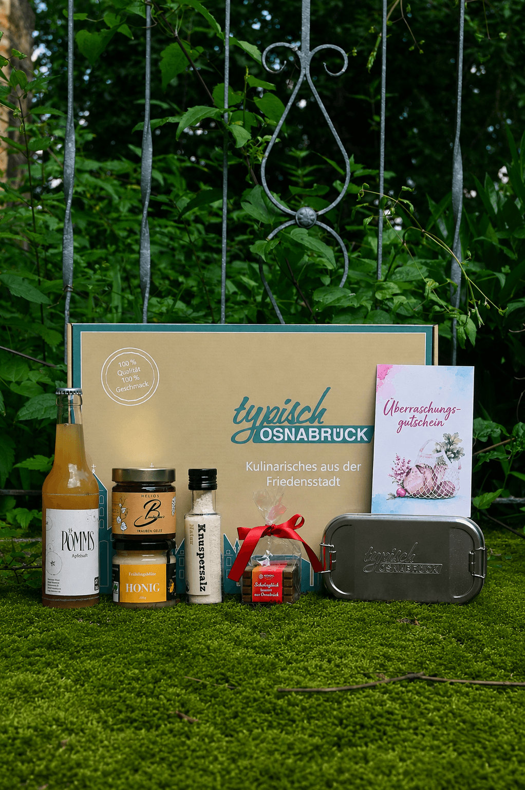 Präsentkarton „Typisch Osnabrück“ vor einem Zaun im Grünen, umringt von Flasche, Gläsern, Salz, Schokolade und Edelstahl-Dose.